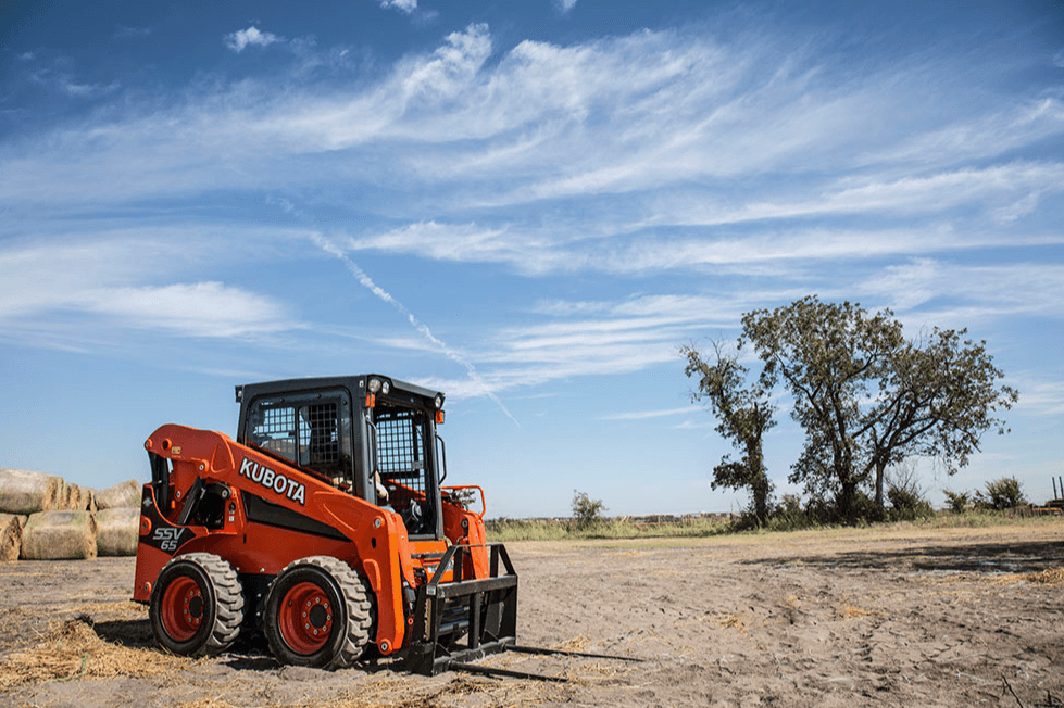 Kubota SSV65 skid steer with pallet forks near hay bales on a farm